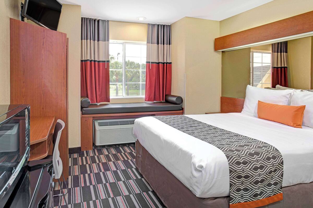 A hotel room with a bed, a desk, a chair, a window with curtains, and a view of the outdoors.
