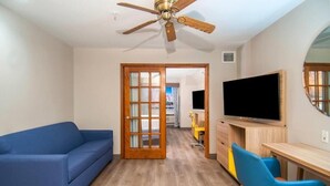 A room with a blue sofa, a wooden desk with a TV, a ceiling fan, and a door leading to another room.