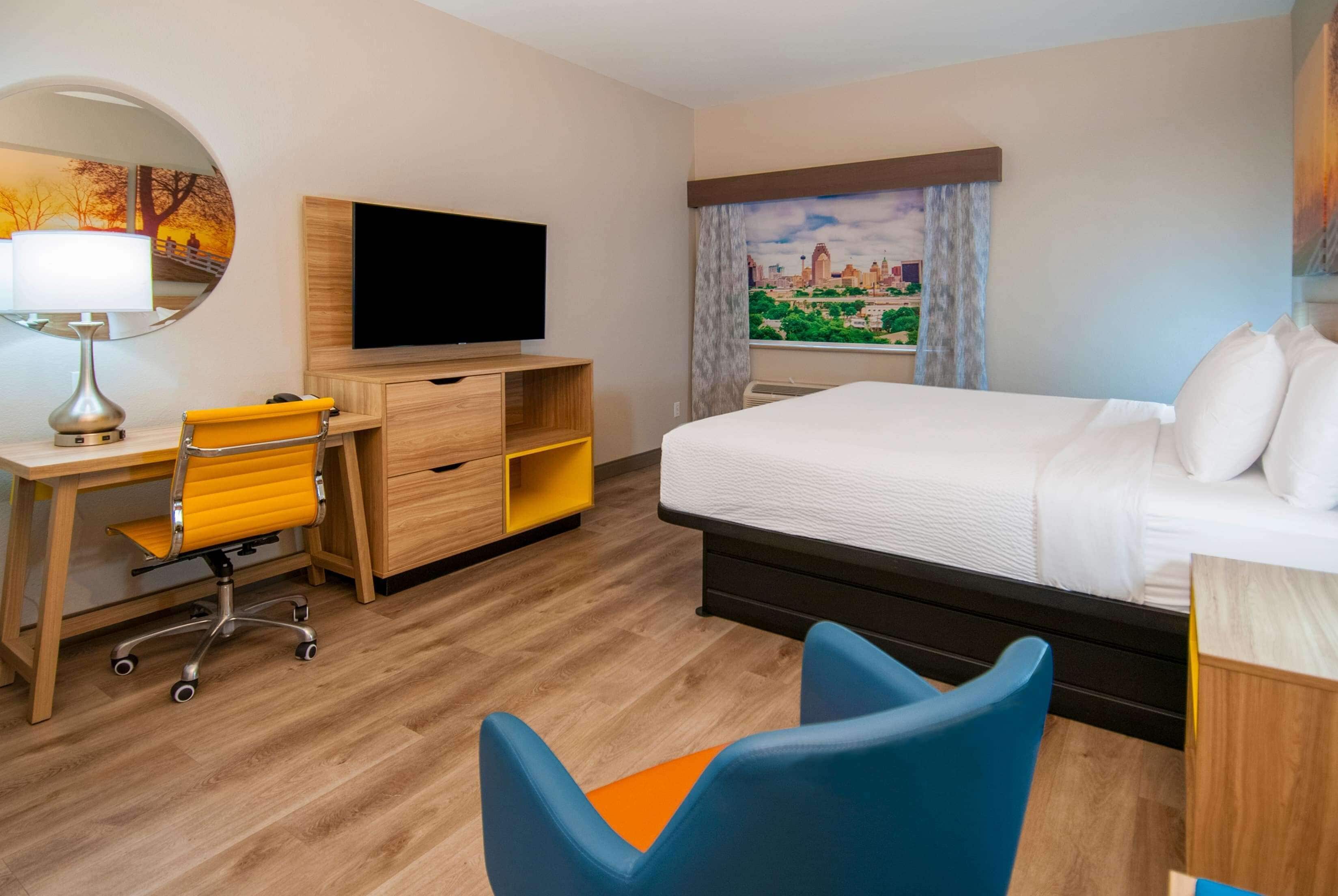 A hotel room with a bed, a desk with a chair, a TV, and a window with a cityscape view.