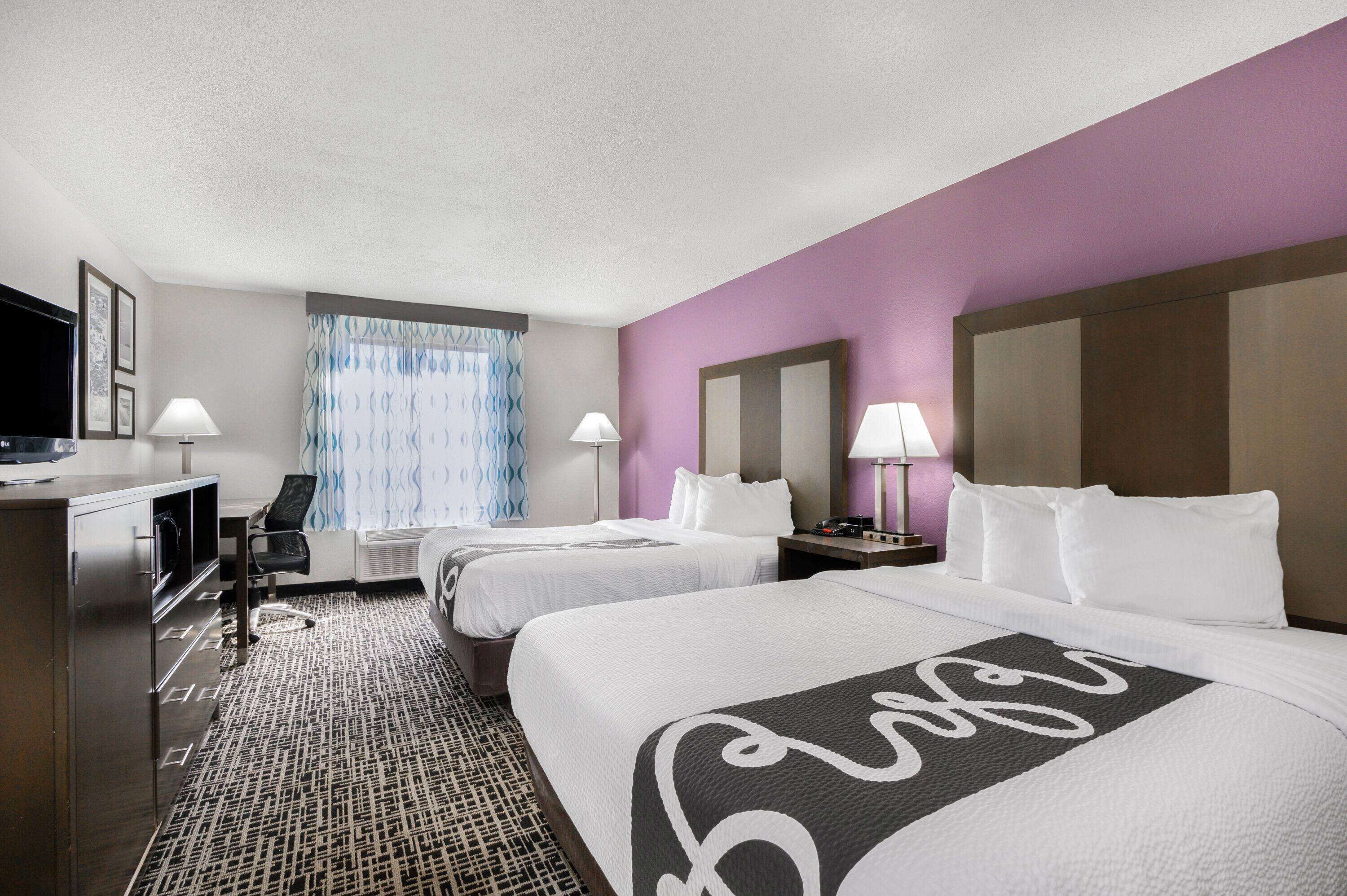A hotel room with two beds, a desk, a TV, and a window with curtains.