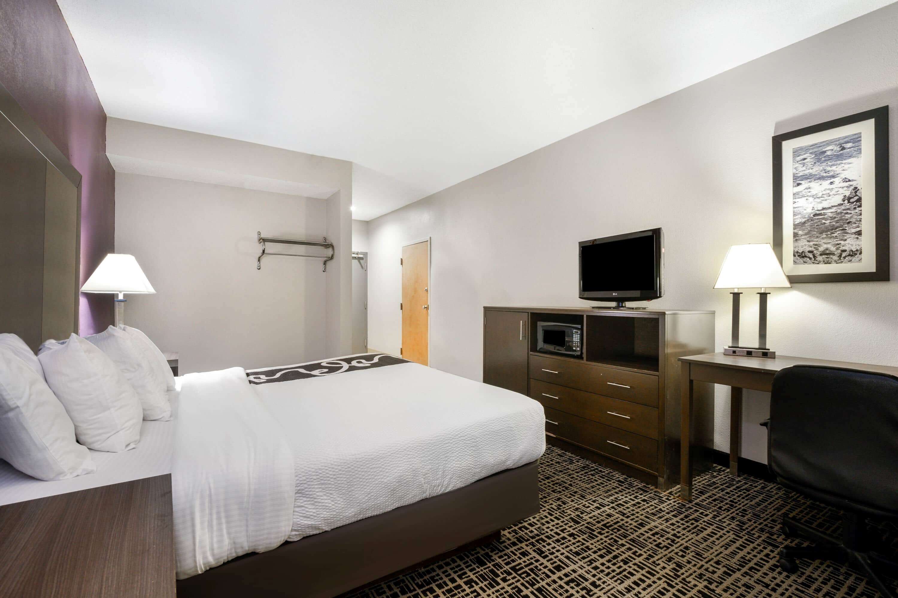 A hotel room with a bed, a desk with a lamp, a television, a microwave, and a framed picture on the wall.
