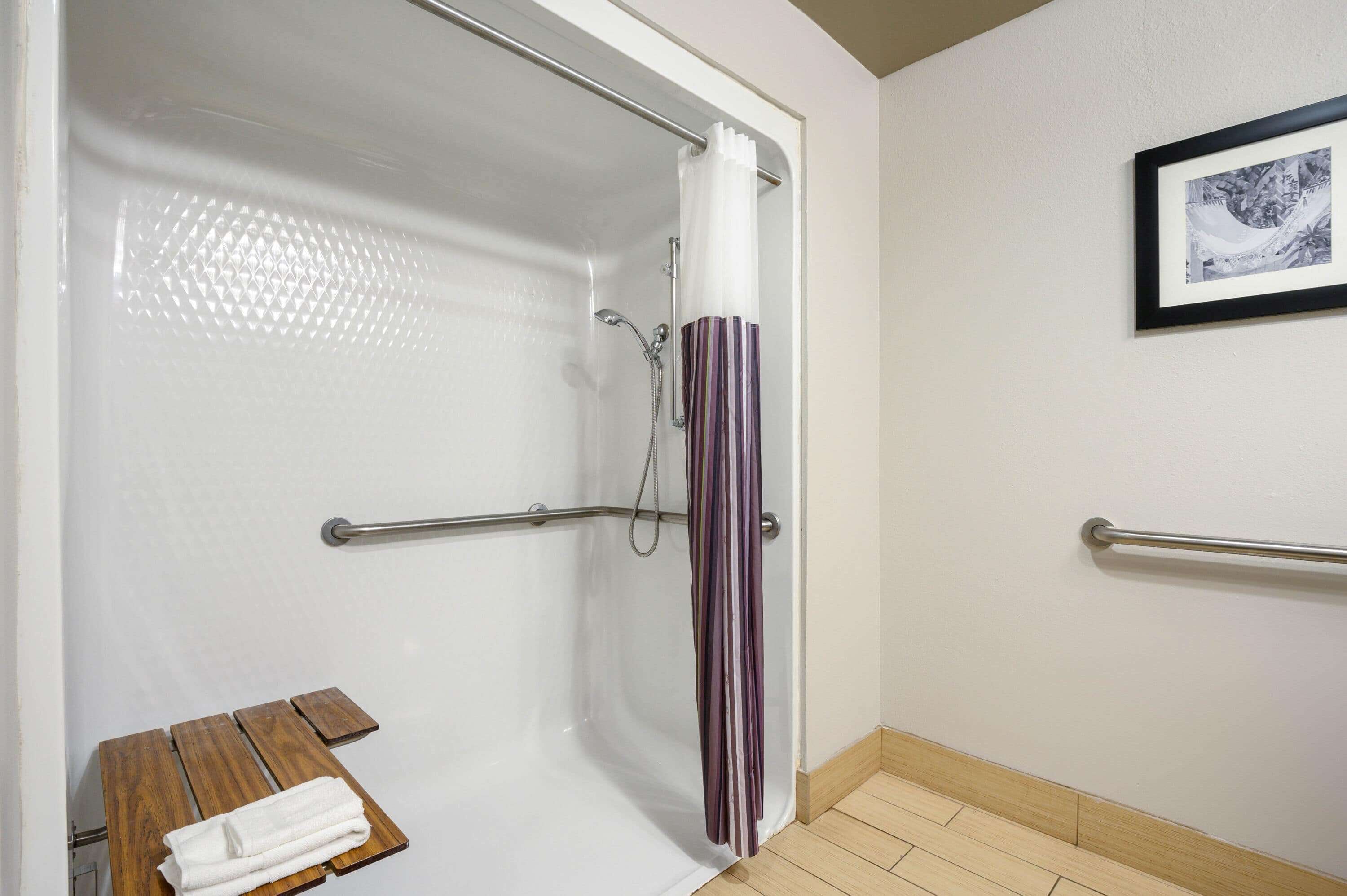 A shower with a glass door, a handrail, and a wooden bench with a towel.
