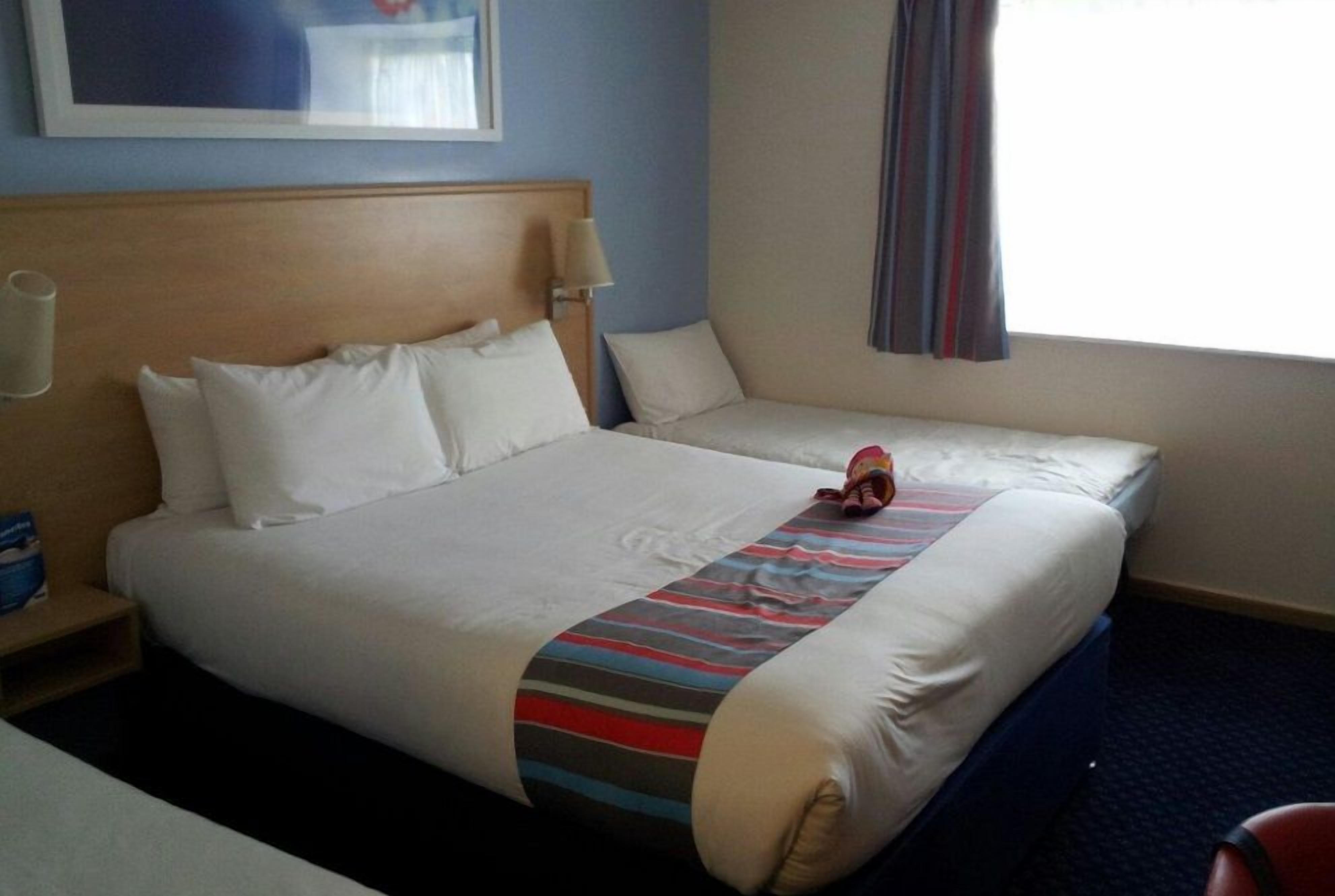 A hotel room with a large bed, a smaller bed, a nightstand, a lamp, and a window with curtains.