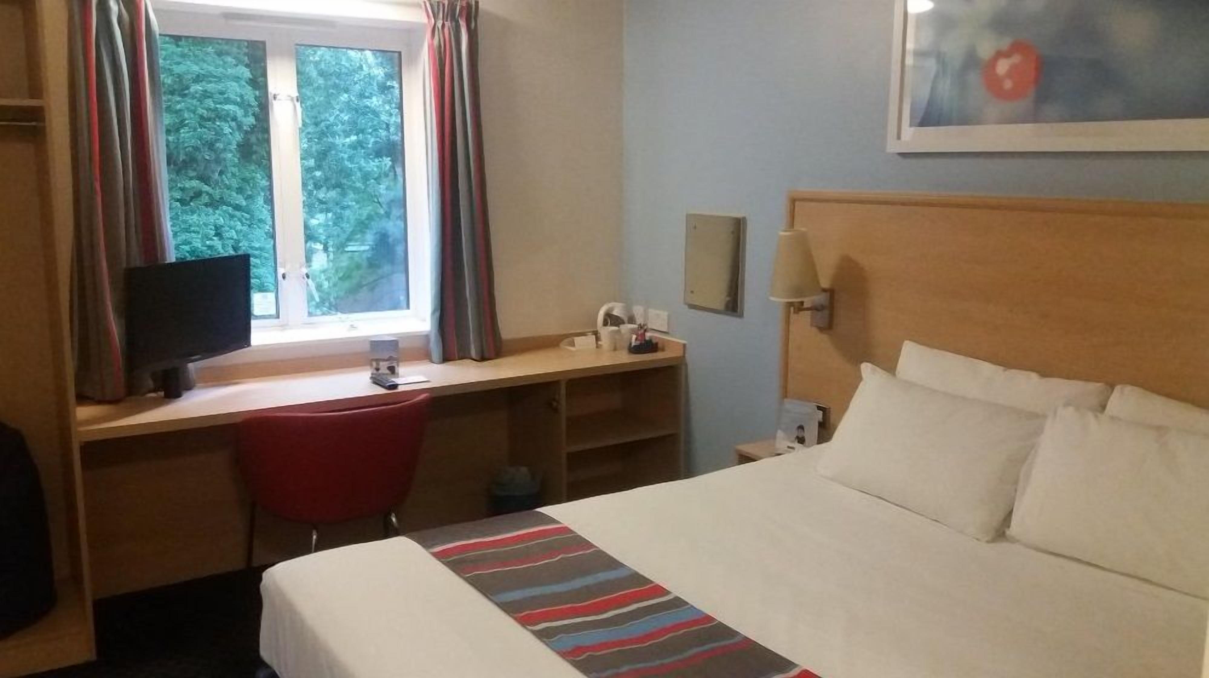 A hotel room with a bed, a desk with a computer, a chair, a window with curtains, and a painting on the wall.
