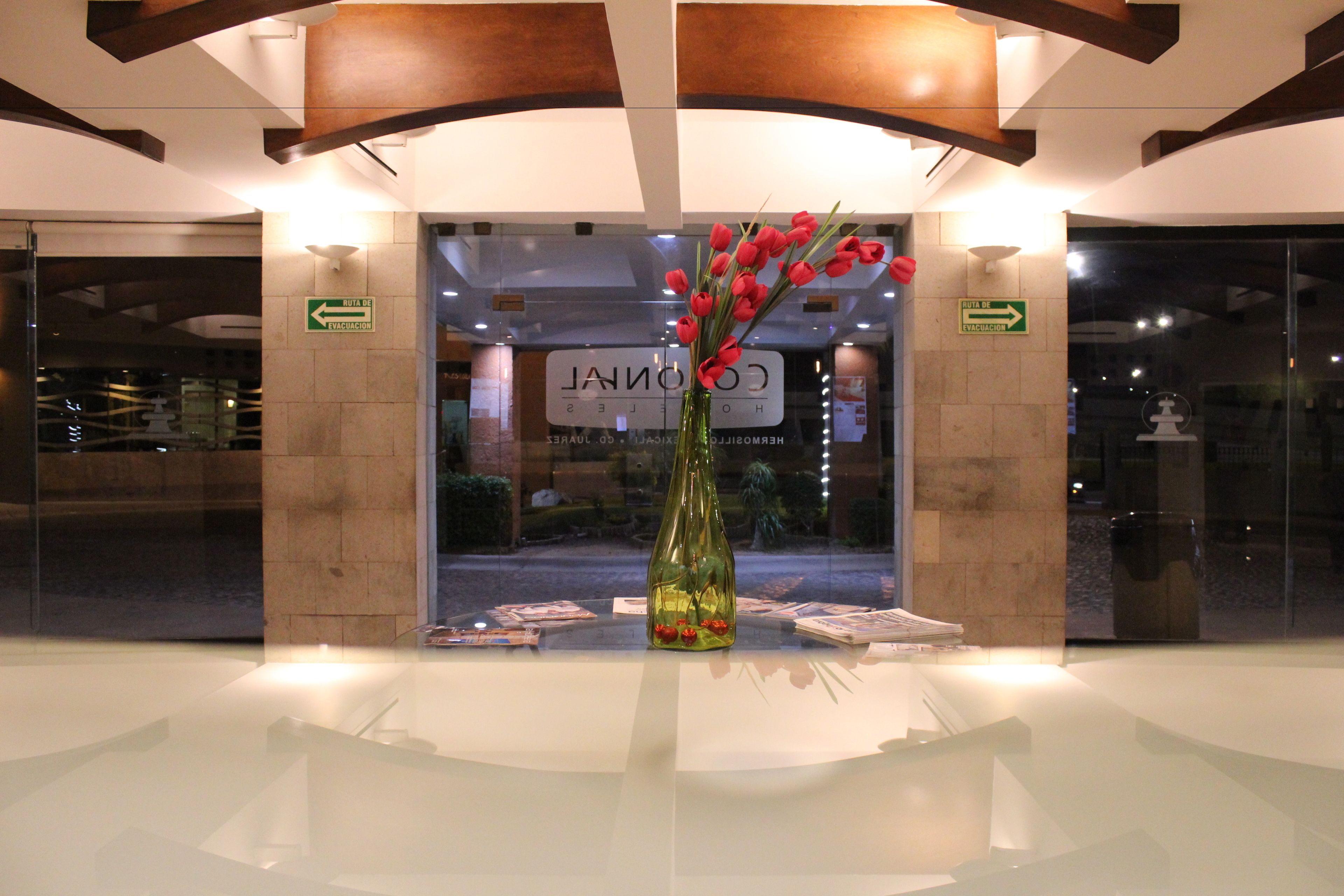 Un área de recepción con puerta de vidrio, un jarrón con flores rojas y señales indicativas de rutas de evacuación.