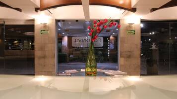 Un área de recepción con puerta de vidrio, un jarrón con flores rojas y señales indicativas de rutas de evacuación.