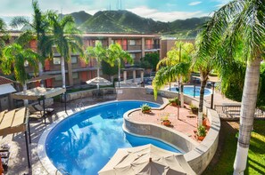 Relax by the resort pool surrounded by palm trees and umbrellas, with a scenic view of rolling hills in the background.