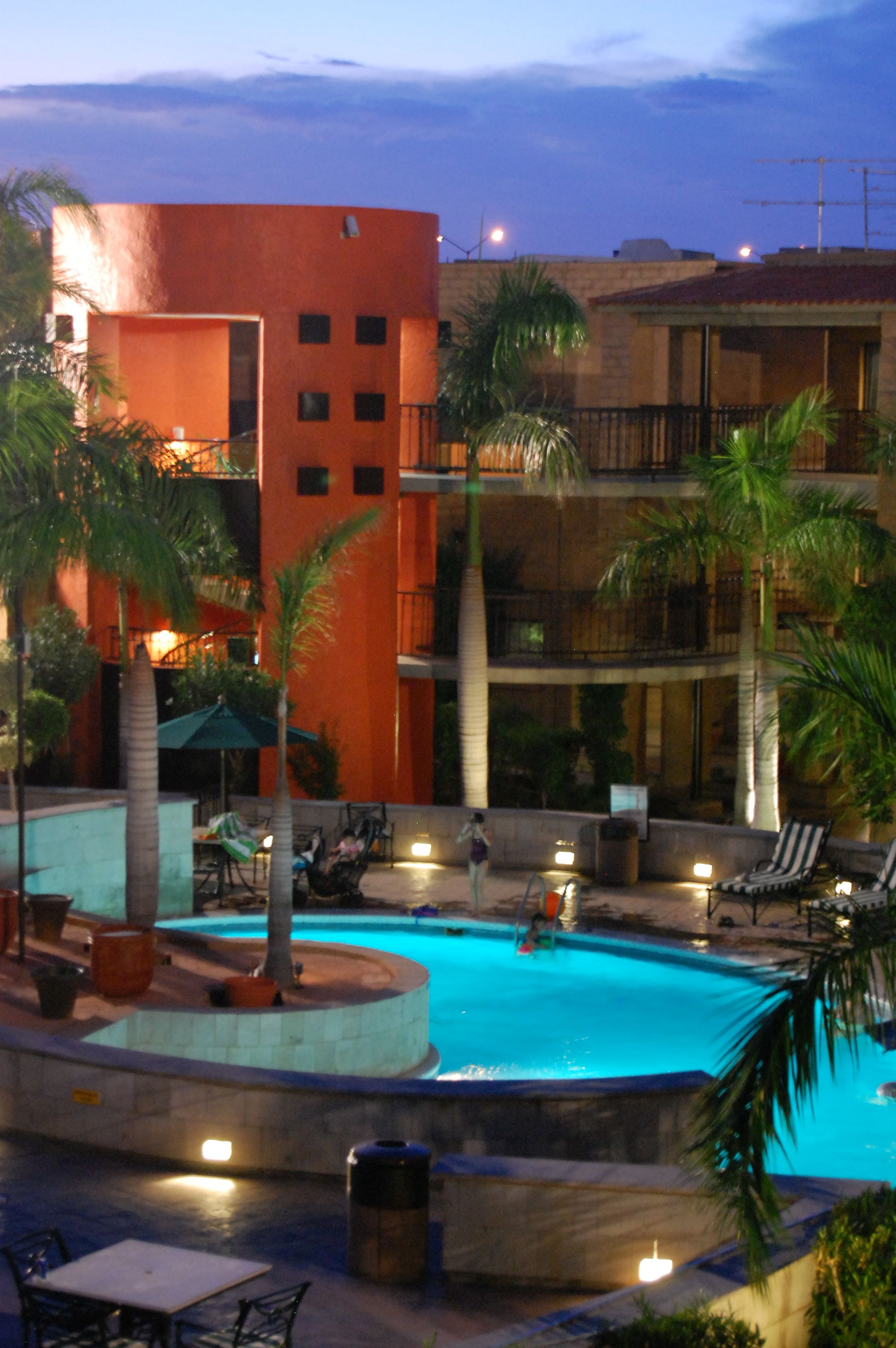 A swimming pool surrounded by palm trees and lounge chairs, with a building featuring balconies and a flat roof in the background.