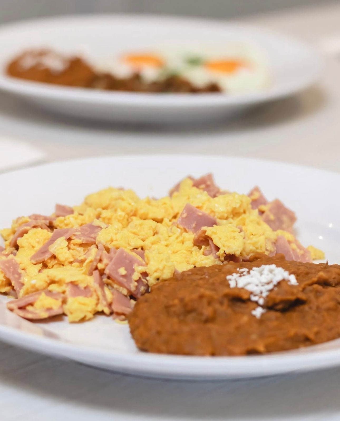 A plate of scrambled eggs with ham and a piece of brown food, possibly a bean dish, on a white plate.