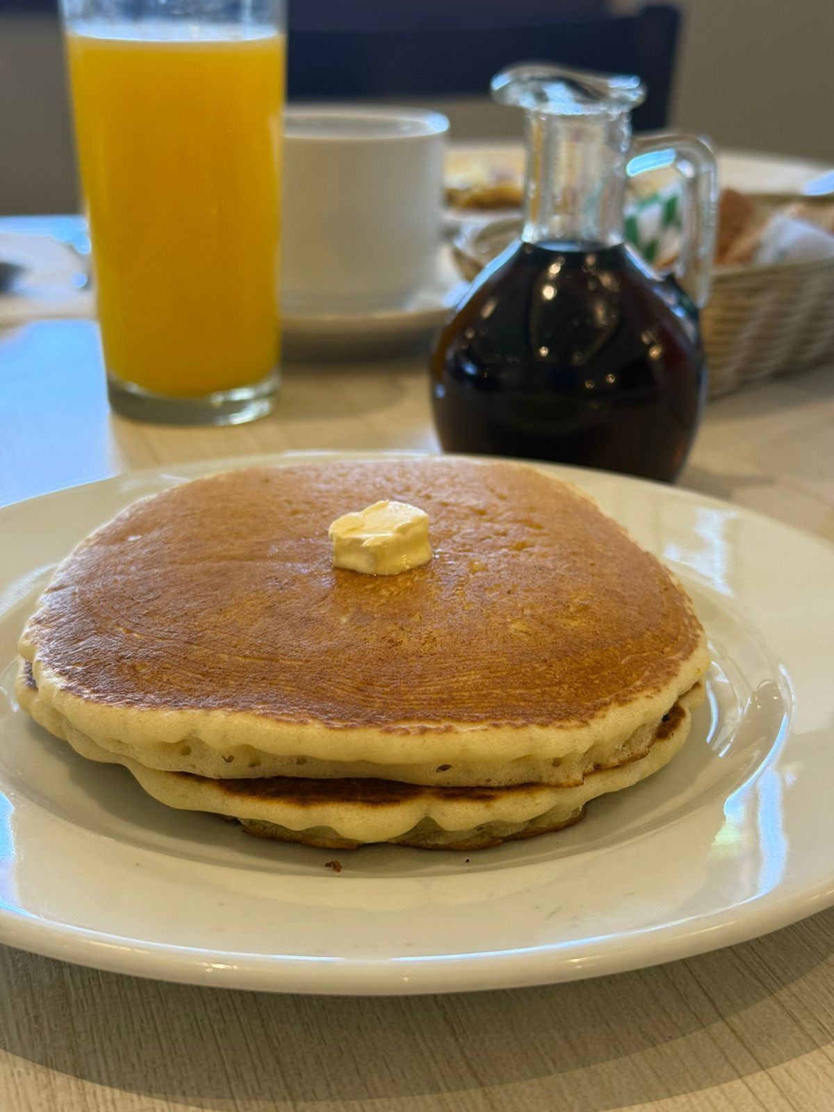 A plate of pancakes with butter on a white plate, a glass of orange juice, and a bottle of syrup on a table.