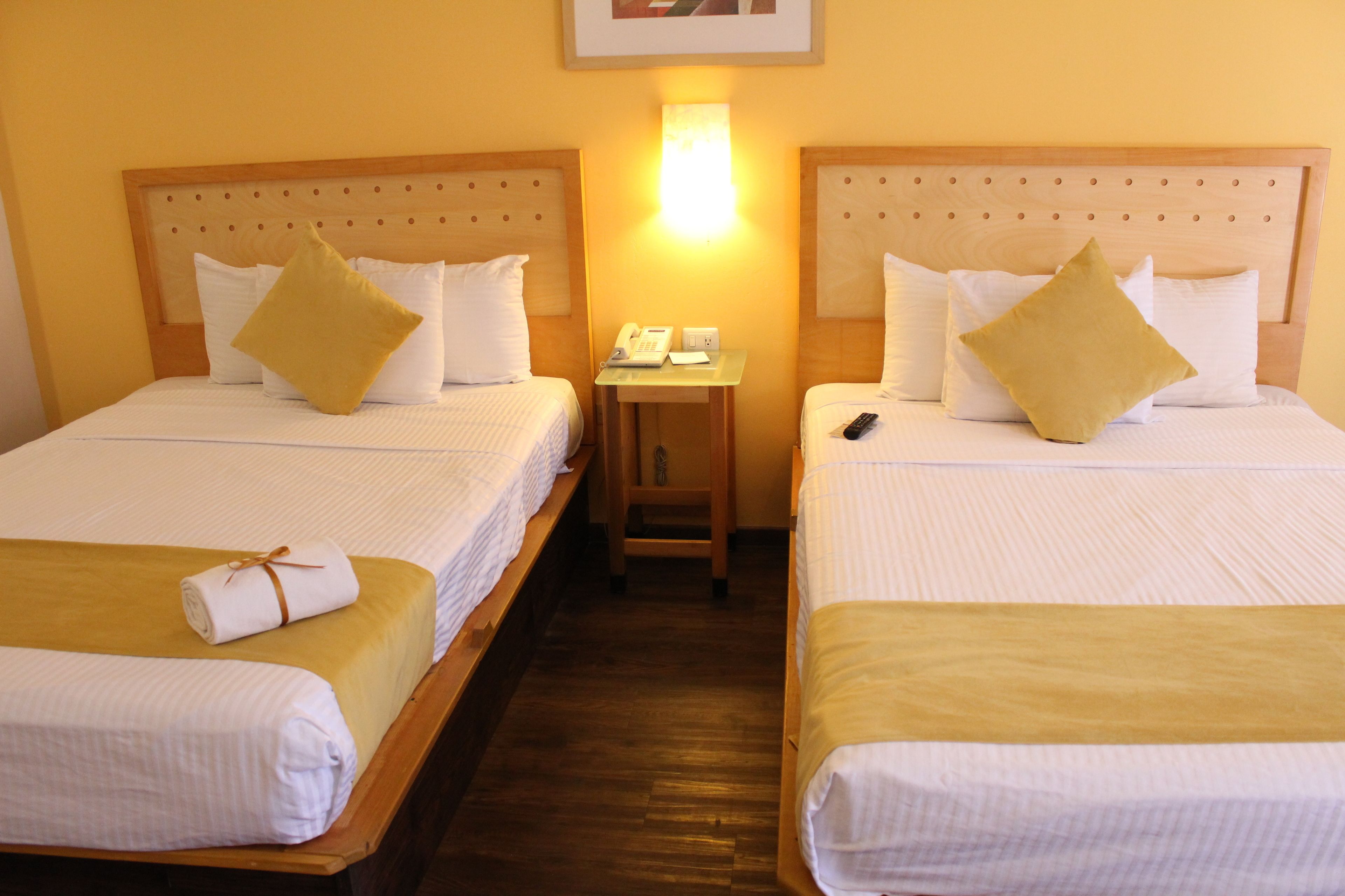 Habitación de hotel con dos camas individuales, cada una con una almohada y una toalla enrollada. Hay una mesita con un teléfono y una lámpara.