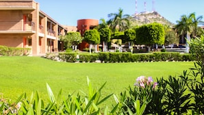 A hotel courtyard with a well-maintained lawn, a building with a brick facade, and a hill with palm trees in the background.