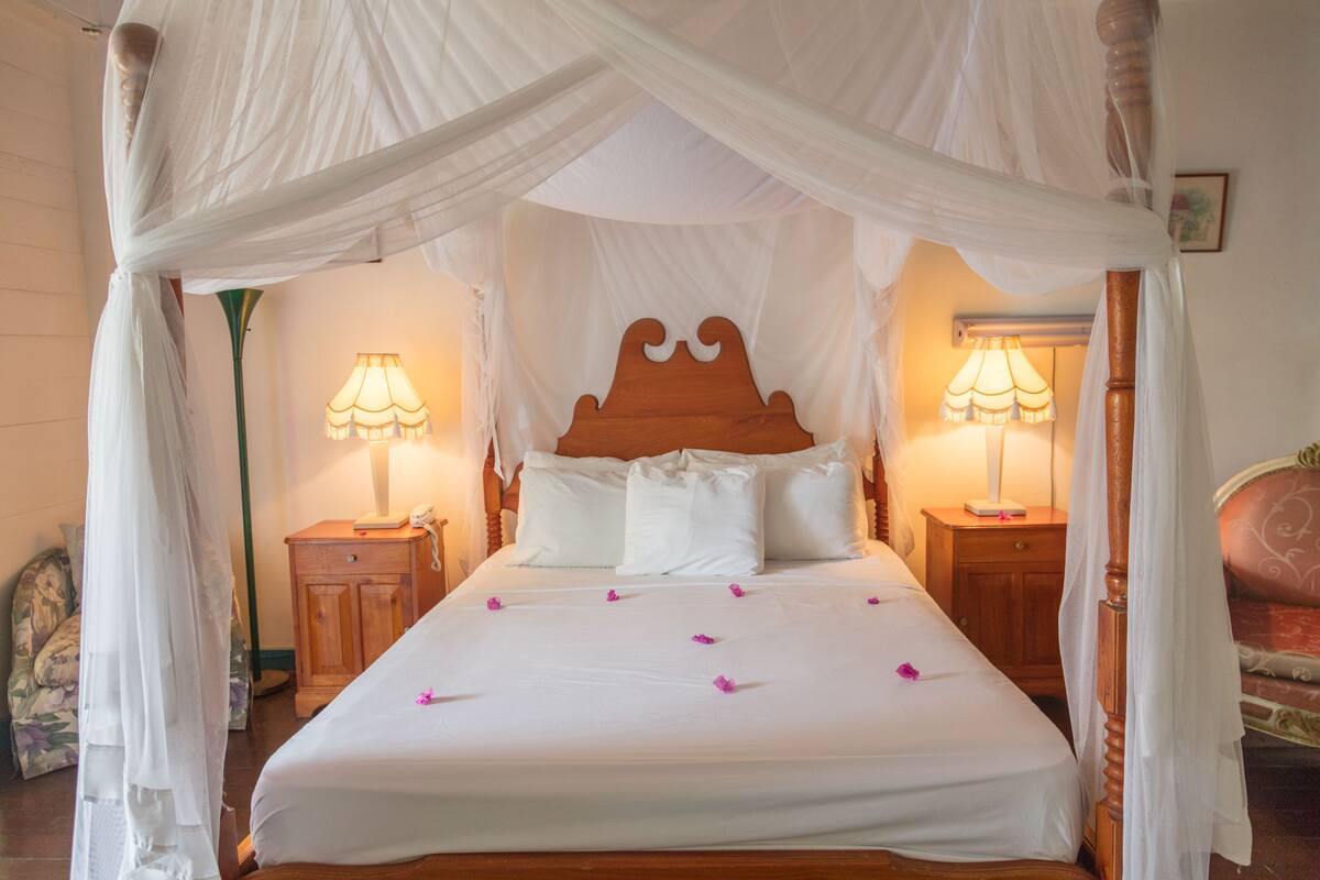 Una cama con dosel y cuatro columnas, con sábanas blancas y pétalos de flores rosas, flanqueada por mesitas de noche de madera con lámparas.