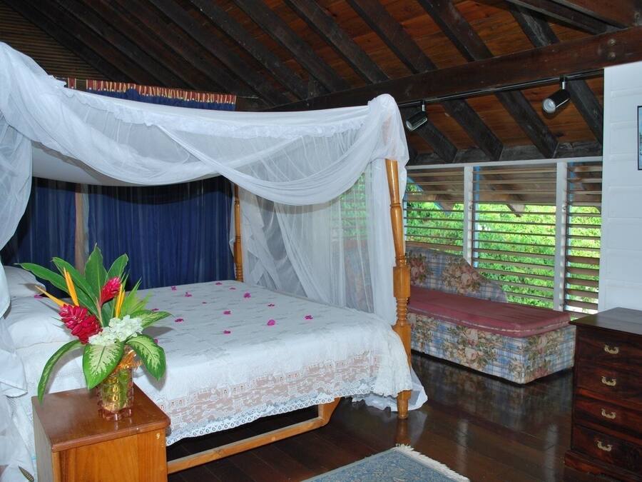A bedroom with a four-poster bed, a bedside table with a vase of flowers, a bench, and a wooden dresser.