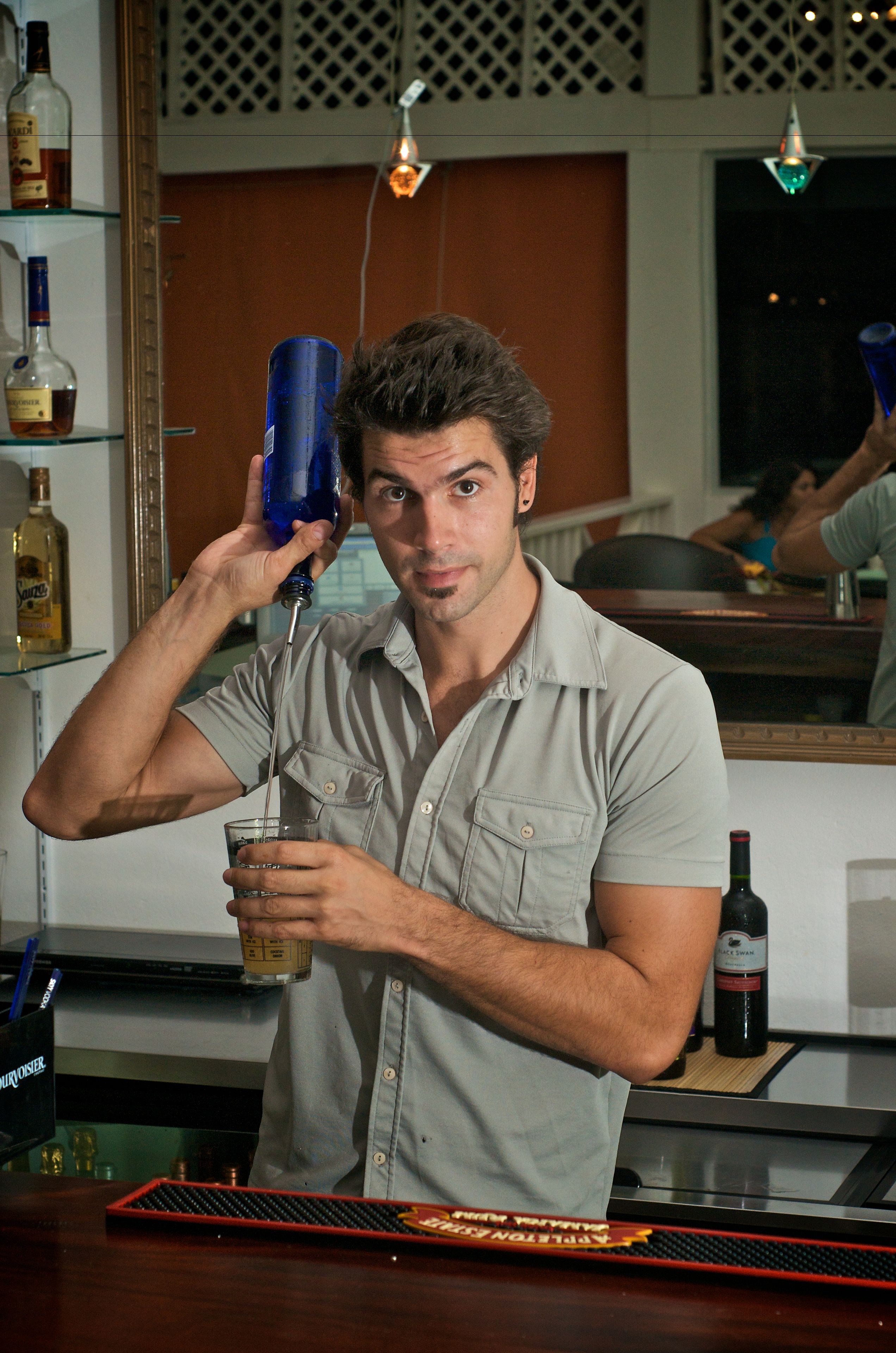 A bar area with a counter, a person pouring a drink, and bottles of liquor on shelves.