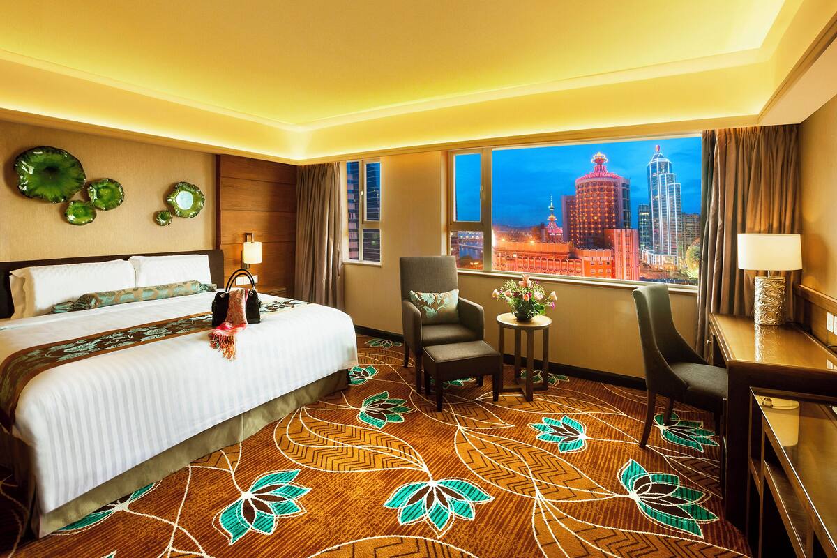 Habitación de hotel con una cama grande, dos sillones, una mesita con un florero y vistas a un paisaje urbano nocturno.