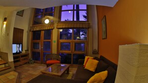 A living room with a sofa, a coffee table, a television, and large windows with a view of palm trees. - Villas El Rancho (Mazatlán)