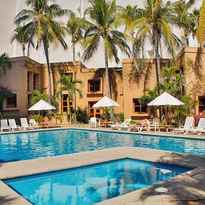 A swimming pool with lounge chairs and umbrellas, surrounded by palm trees and a building with multiple windows.