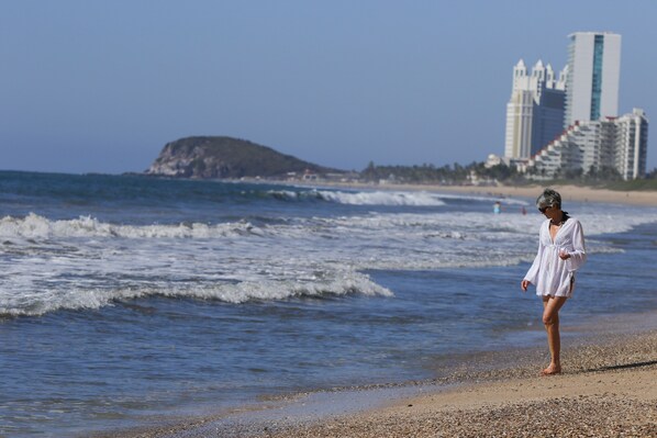 A beach with a person walking along the shore, waves crashing onto the beach, and a distant view of a cityscape with tall buildings. - Villas El Rancho (Mazatlán)