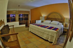 A bedroom with a large bed, two bedside tables with lamps, a wooden chair, and a window with a view. - Villas El Rancho (Mazatlán)