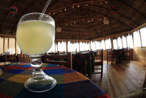 A glass of a light-colored beverage on a table with a colorful tablecloth, set in a room with wooden floors and a thatched roof, featuring multiple tables and chairs. - Villas El Rancho (Mazatlán)