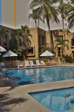 A pool area with lounge chairs and umbrellas, surrounded by palm trees and a building with a yellow stripe.