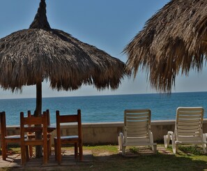 A beachside seating area with wooden and plastic chairs, a thatched umbrella, and a view of the ocean.