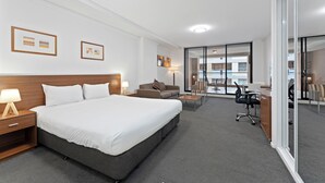 A hotel room with a large bed, a desk, a sofa, and a glass partition. - APX Darling Harbour (Sydney)