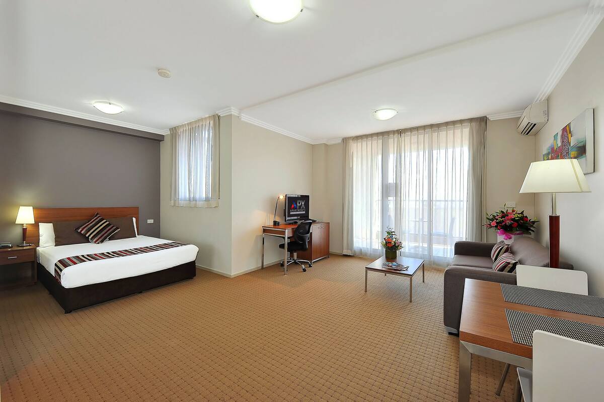 Quarto de hotel com cama, escrivaninha, cadeira e pequena mesa com abajur.