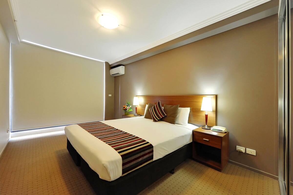 Quarto de hotel com uma cama grande, mesa de cabeceira com abajur e ar condicionado fixado na parede.