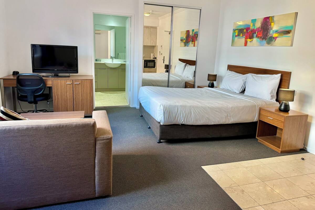 Quarto de hotel com uma cama, mesas de cabeceira, uma escrivaninha com cadeira, televisão e banheiro visível por uma porta aberta.