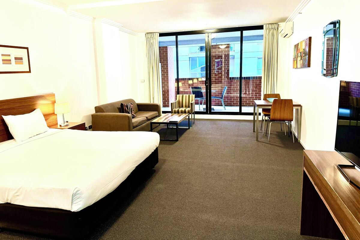 Quarto de hotel com cama, sofá, mesa de centro, escrivaninha e televisão.
