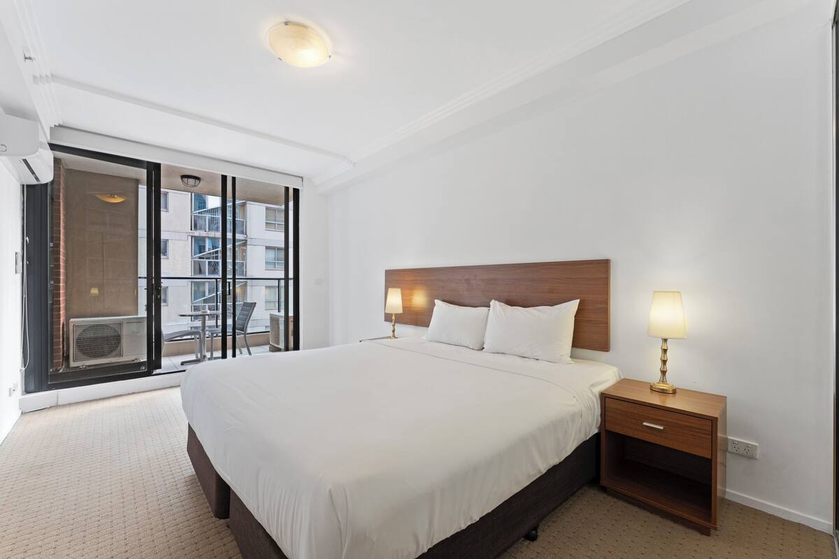 Quarto de hotel com uma cama grande, mesa de cabeceira com abajur e porta de vidro deslizante para uma varanda externa.
