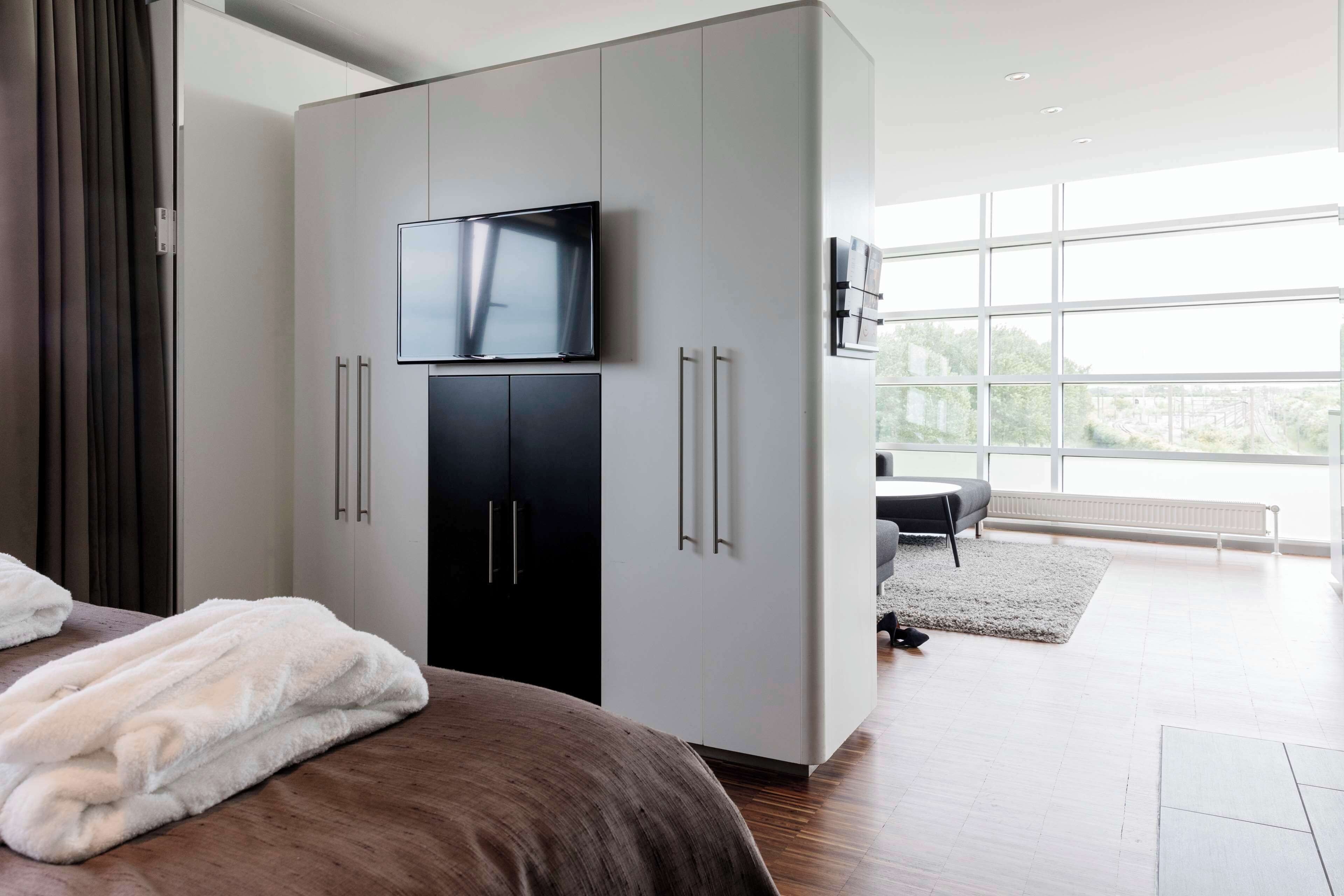 Quarto moderno com cama grande, televisão de tela plana montada sobre um guarda-roupa preto, e vista para o exterior por grandes janelas.