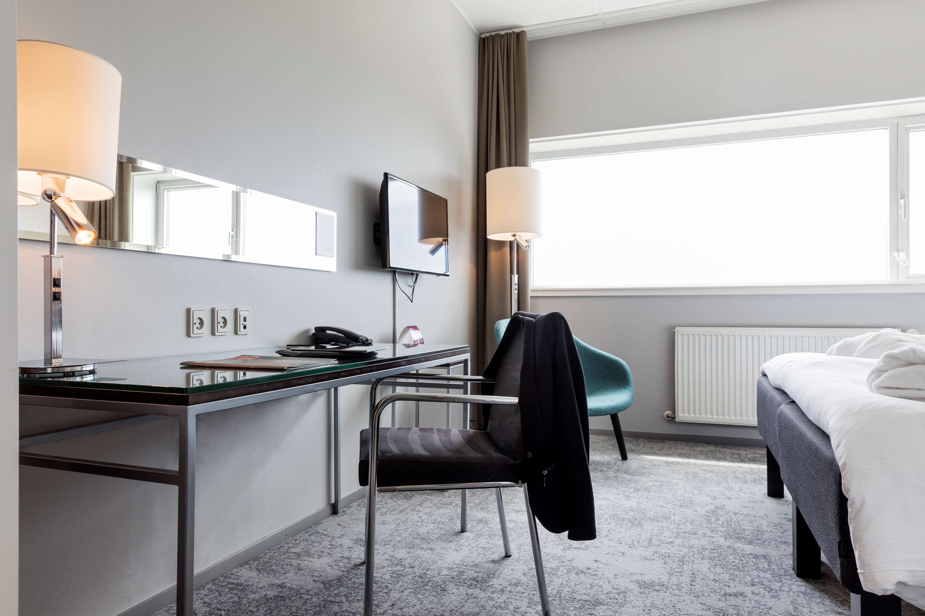 Habitación de hotel con escritorio, silla, cama y ventanas amplias.