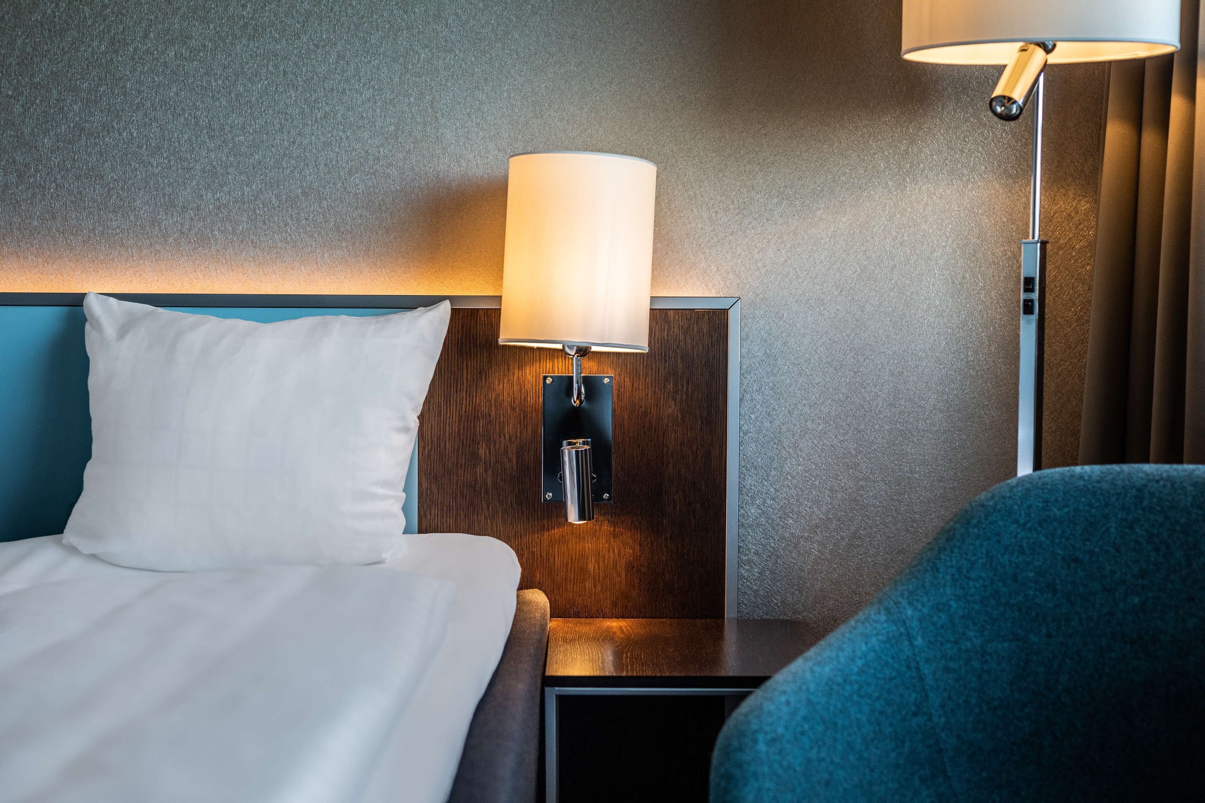 A neatly made bed with a white pillow, a wooden nightstand with a lamp, and a blue chair.