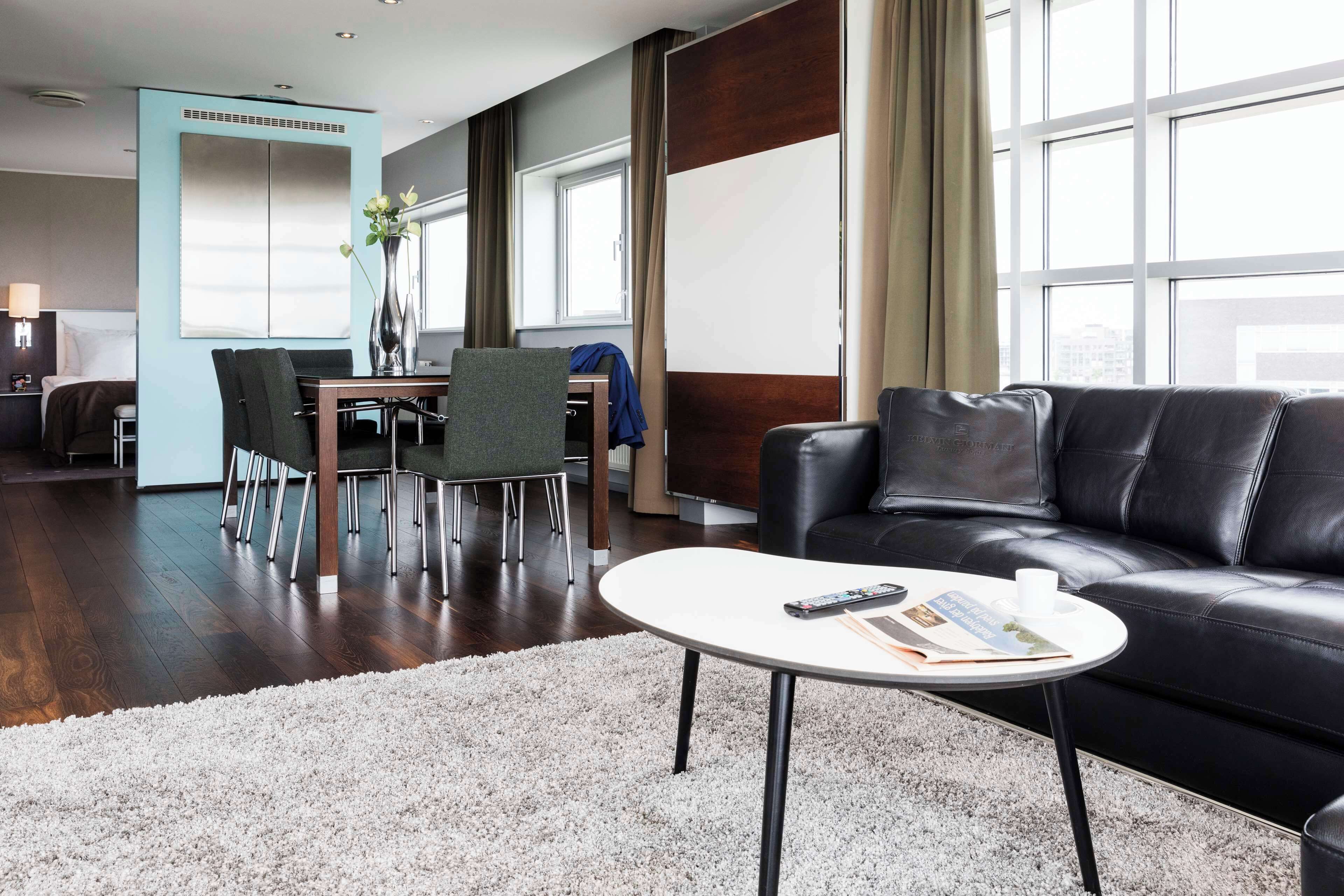 Une chambre d’hôtel moderne avec un coin repas, un canapé en cuir noir et une table basse ronde.