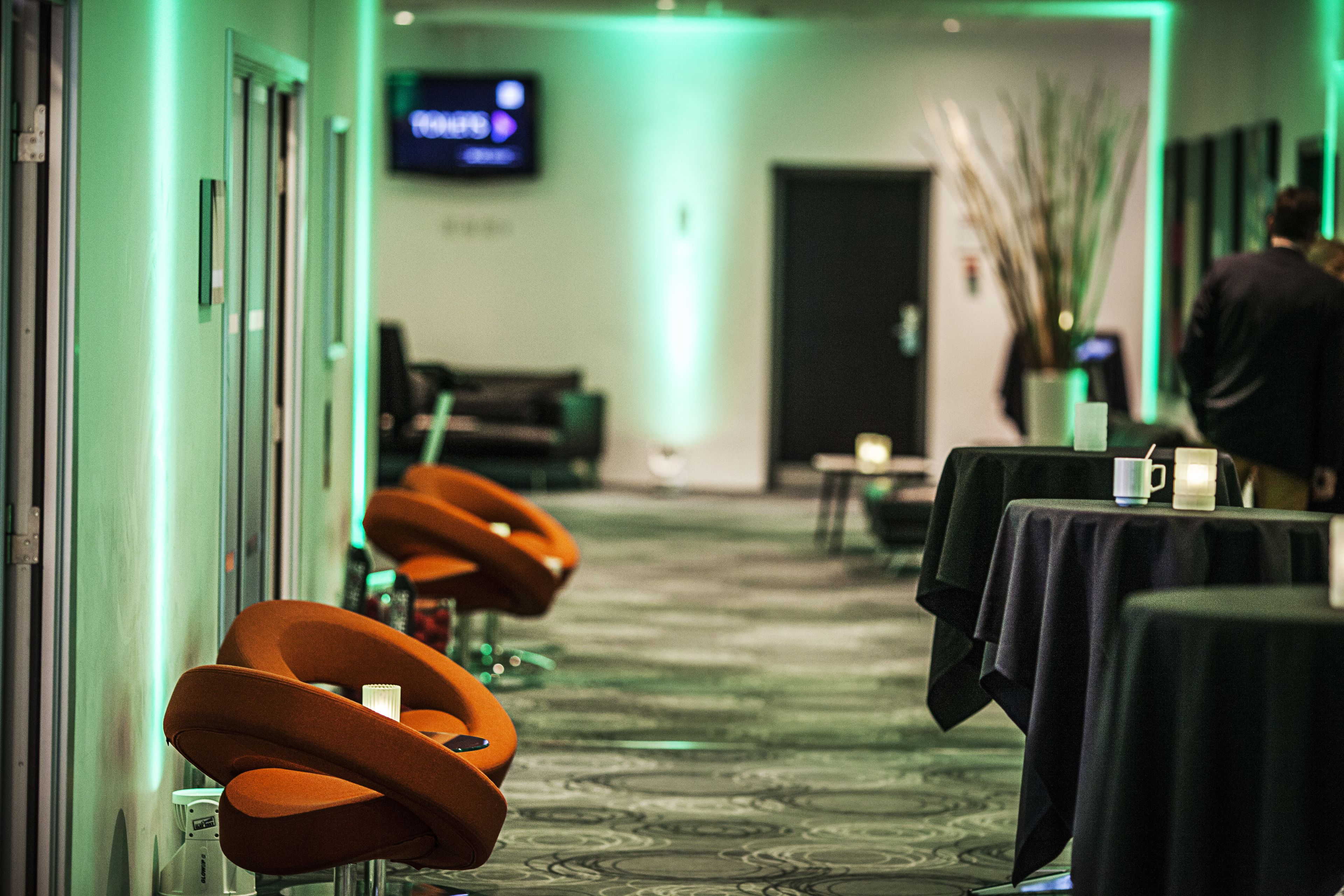 A hotel corridor with a green-lit reception area, a TV screen, and a row of orange chairs. Tables with black tablecloths and a vase of branches are visible.