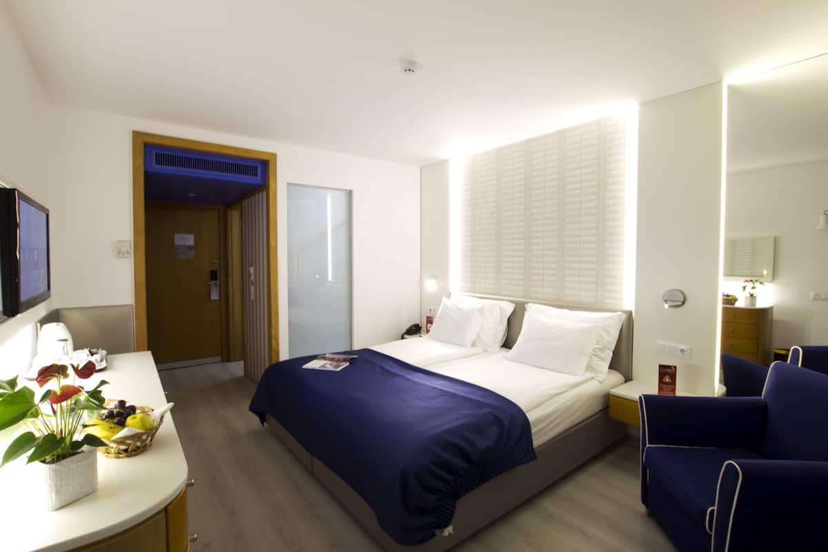 A hotel room with a bed, bedside table, chair, and a small desk with a lamp.