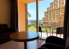 Habitación de hotel con balcón, una mesa redonda y dos sillas. El balcón tiene vistas a una piscina y un edificio con varios balcones.