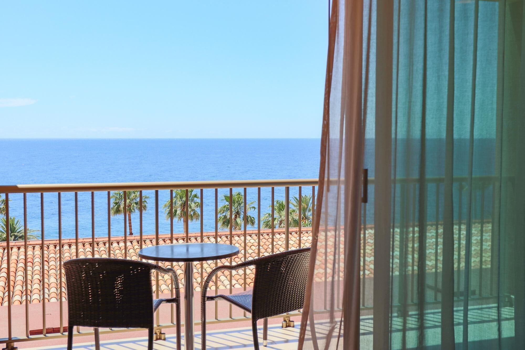 A balcony with two wicker chairs and a small round table, overlooking a beach with palm trees and the ocean.