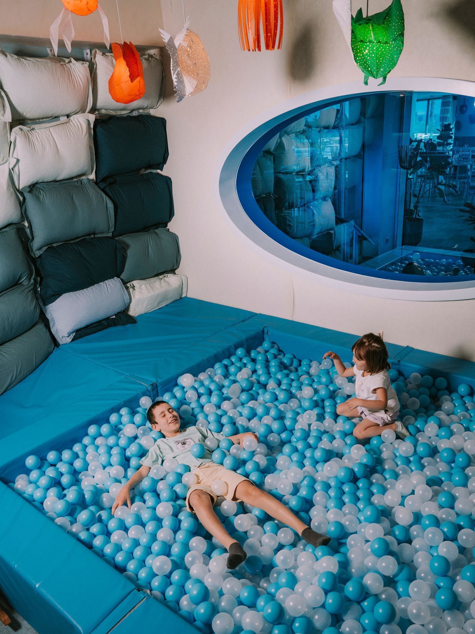 Ein Zimmer mit einer blauen, gepolsterten Spielzone voller blauer und weißer Bälle, einer Sitzecke mit Kissen und einem runden Fenster mit Blick auf eine Stadtansicht.