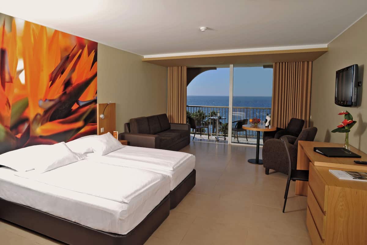 A hotel room with a bed, a desk, a chair, a TV, and a balcony with a view of the sea.