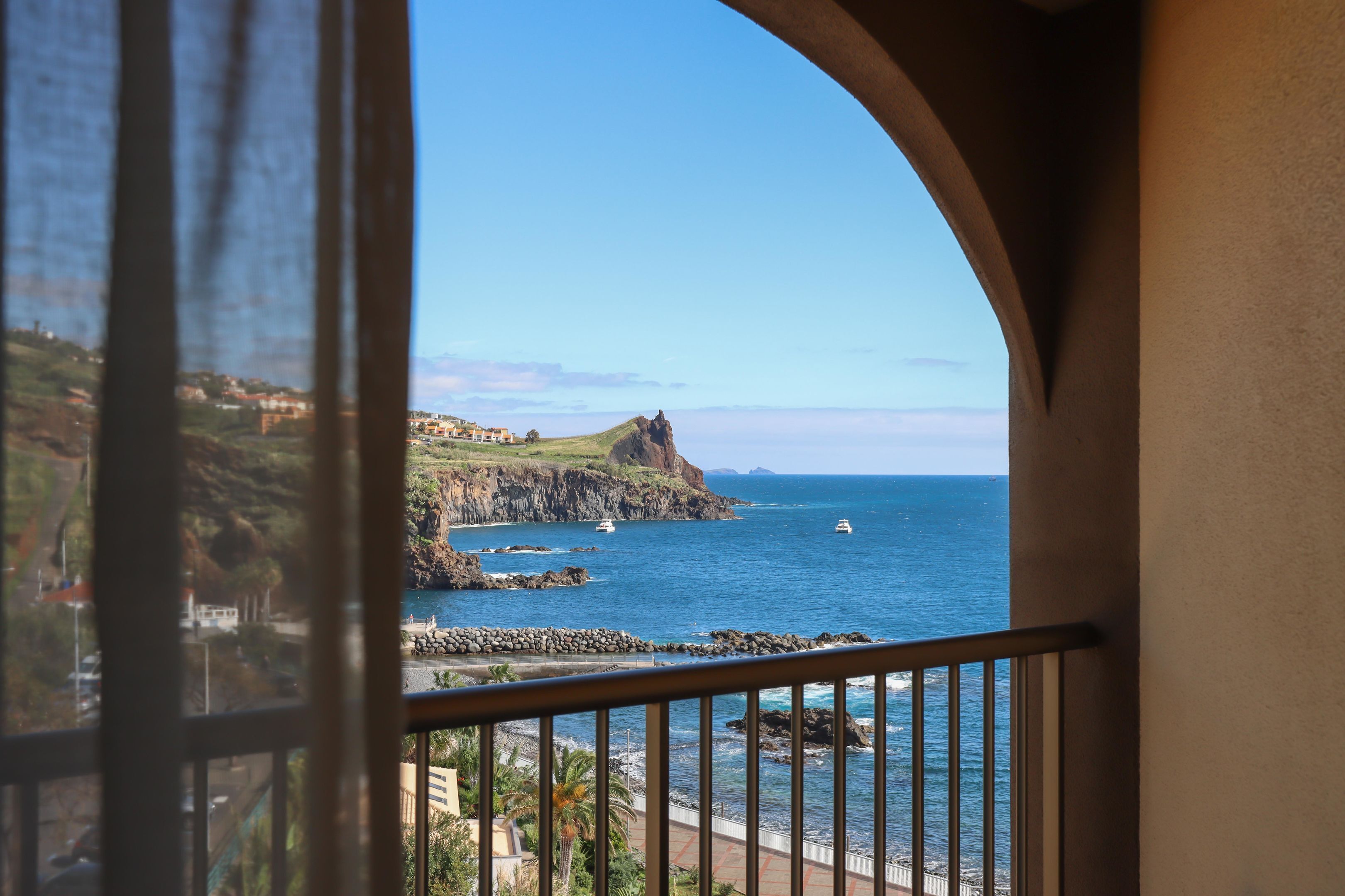 Vista costera desde un balcón con barandilla de piedra, costa rocosa y cielo despejado.