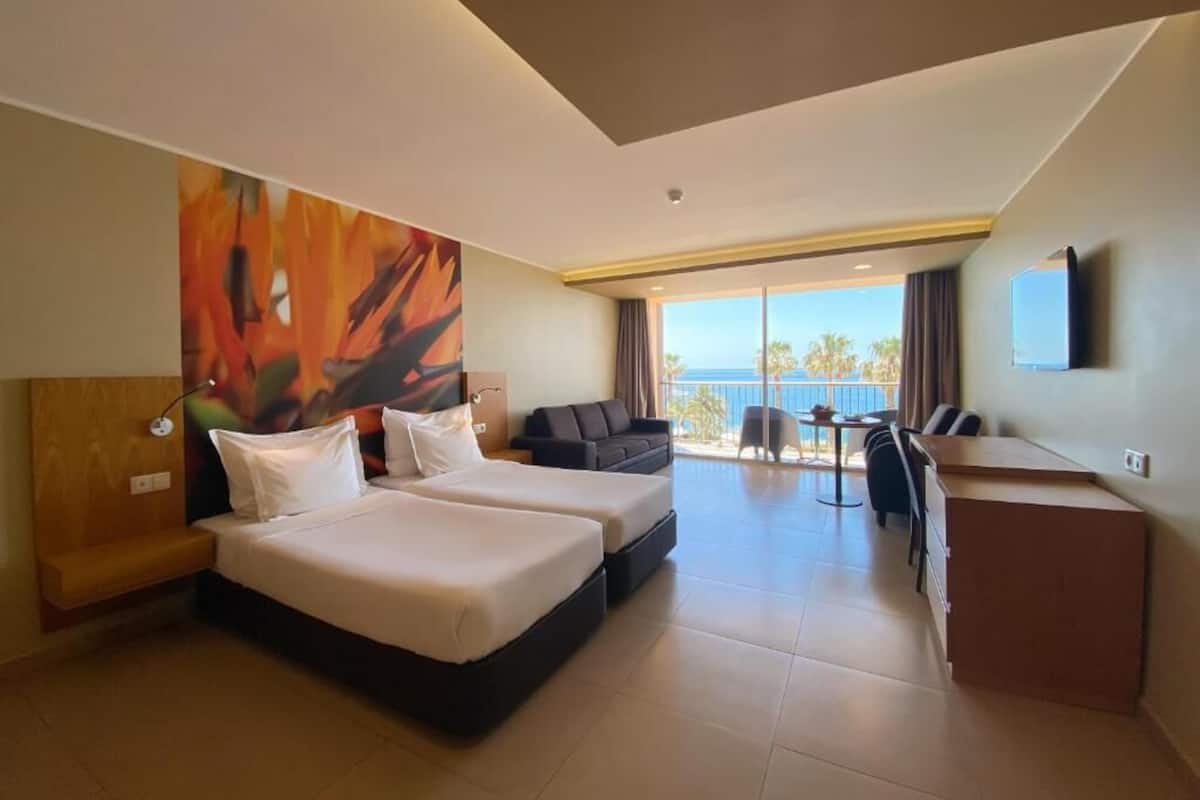 A hotel room with two beds, a desk, a chair, a sofa, a TV, and a balcony with a view.
