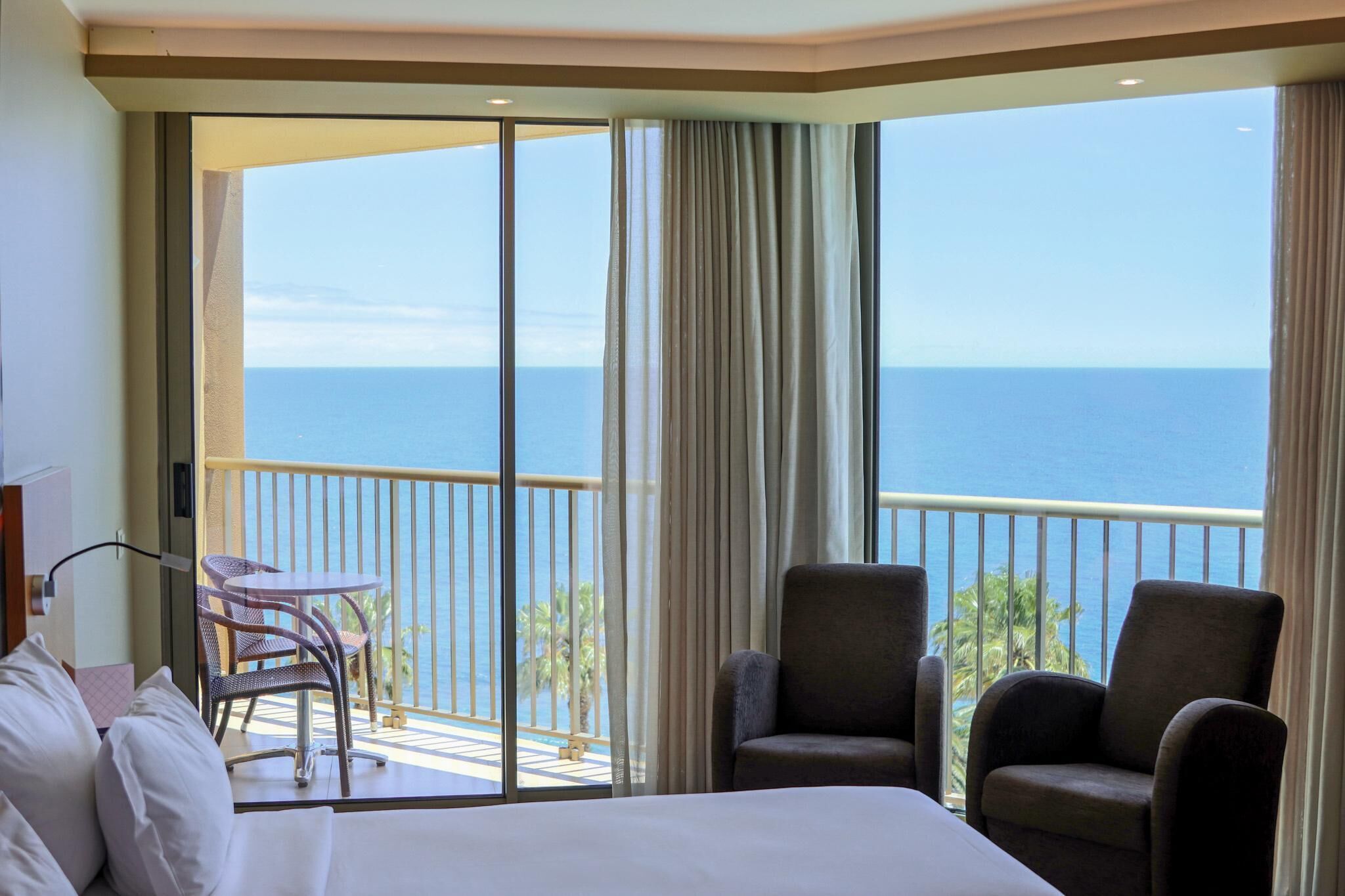 Ein Hotelzimmer mit einem großen Bett, zwei Sesseln, einem kleinen Tisch und einem Balkon mit Meerblick.