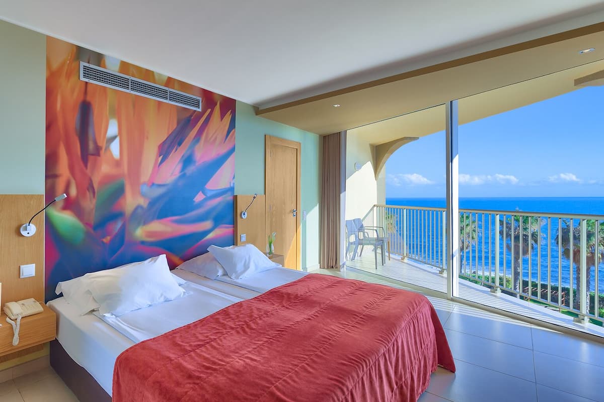 A hotel room with a large bed, a view of the ocean, and a balcony with a railing.