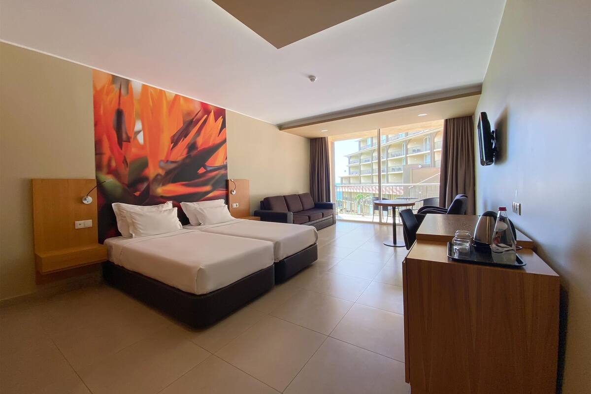 A hotel room with a bed, a sofa, a desk, and a large window offering a view of the building's exterior.