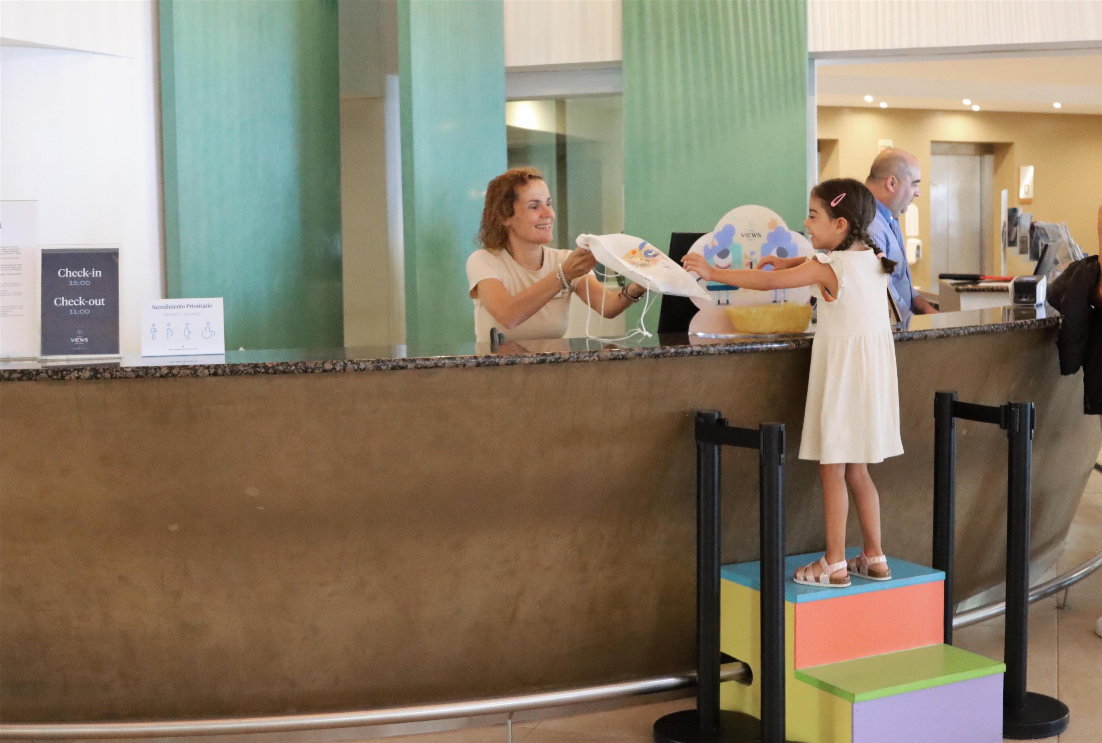 A hotel reception area with a check-in counter, a sign indicating check-in times, and a child standing on a colorful step.