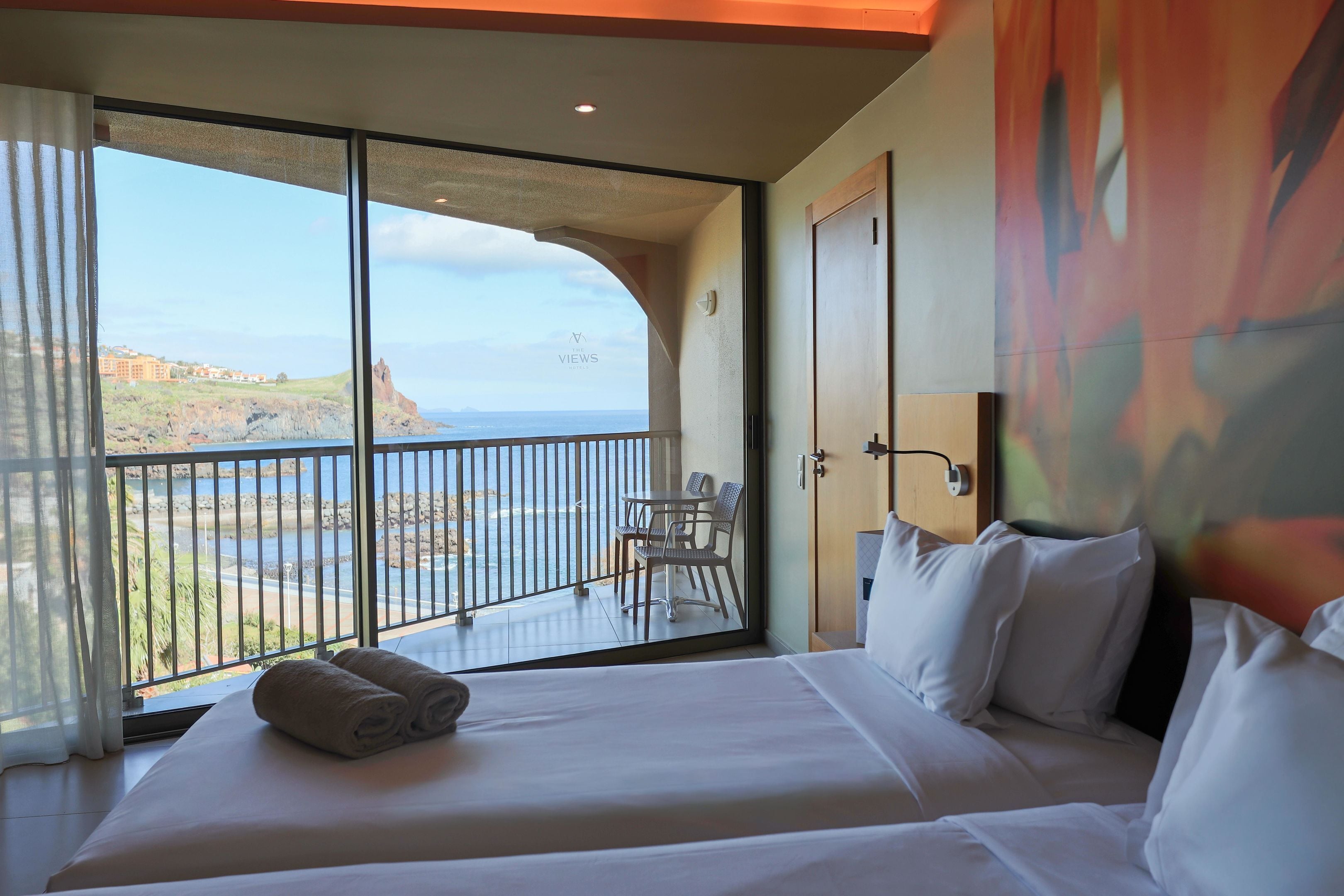 Habitación de hotel con cama, vista al mar, balcón con mesa y sillas, y un cuadro en la pared.